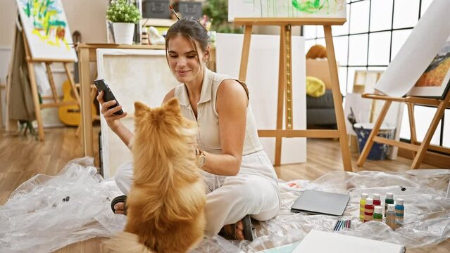 Charming young hispanic woman artist, smiling while sitting on studio floor, confidently texting amidst painting session with pet dog.