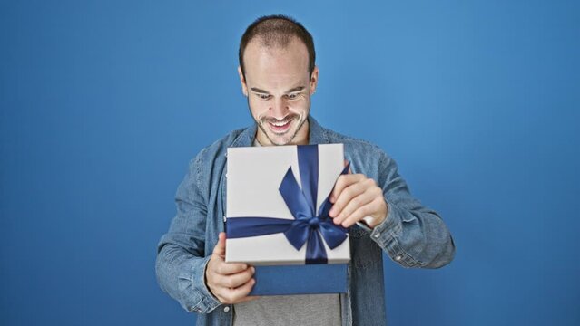A bald, bearded man expressing happiness while holding a blue gift box against an isolated blue background outdoors.