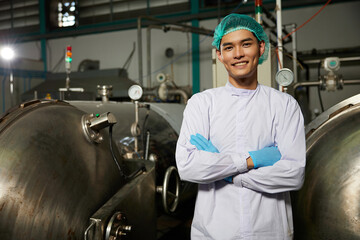 young worker smiling and folded arms pose beside machine control panel or wheel of valve heavy pipe in the factory