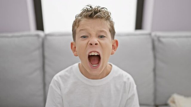 Adorable blond boy sitting on sofa at home, expressing loud frustration and anger