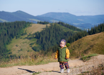 Cute boy walking in mountains at sunny day. Male child in khaki vest looking straight to camera and standing on background of Carpathian mountains.