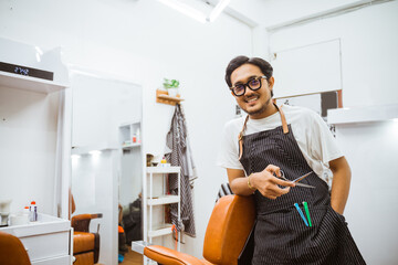 asian male hairdresser holding scissor and leaning on barber chair with big smile on his face - obrazy, fototapety, plakaty