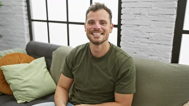 Cheery hispanic young man looking positive and confident sitting on sofa in home interior, his bearded face portraying a healthy, tooth-filled smiling expression of pure delight and success