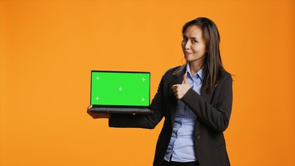 Fototapeta na wymiar Business person holds laptop with greenscreen on camera, showing blank copyspace display on portable pc. Asian woman in formal suit presents computer with chromakey screen in studio.