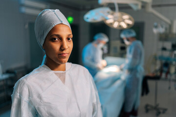 Closeup portrait of African-American female nurse in surgical uniform standing posing looking at camera in operating theater. Multiethnic team of surgeons performing operation on blurred background.