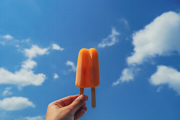 Hand holding an orange popsicle with the sea in the blue sky background. Delicious orange stick ice cream. Refreshing fruit ice creams against a beach scene