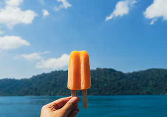 Hand holding an orange popsicle with the sea in the blue sky background. Delicious orange stick ice cream. Refreshing fruit ice creams against a beach scene