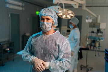 Portrait of confident male doctor in surgical mask and hat in operating room. Practitioner standing posing looking at camera in hospital, team of diverse surgeons performing operation on background