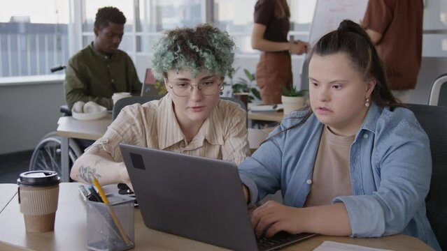 Waist up of young Caucasian woman with down syndrome learning to use laptop and female co-worker helping her during work day in contemporary open space office