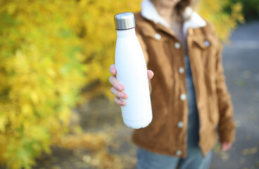 Woman holding a white thermos bottle against the background of autumn trees - obrazy, fototapety, plakaty