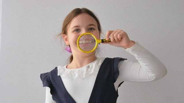 A positive curious schoolgirl in uniform with a wide smile looks at the camera holding a magnifying glass in front of her mouth, a wide smile, teeth enlarged. The concept of dentistry.