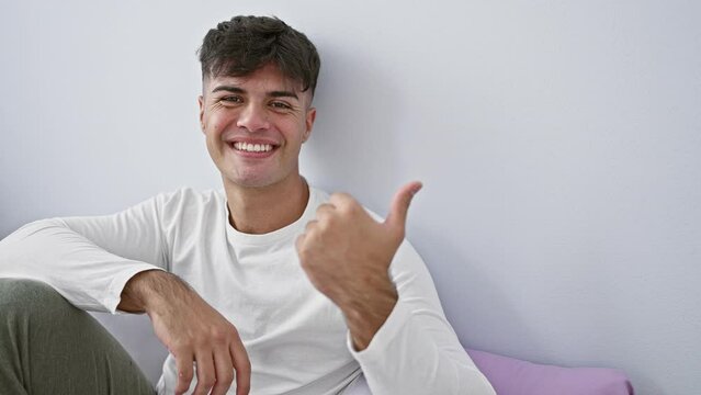Cheerful young hispanic man chillin' in pyjamas, delightfully pointing with thumb, lying comfy on bed in cozy bedroom, playfully looking to the side. perfect space for your ad!