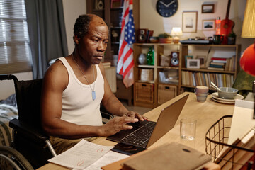 African american man with disability in homewear sitting at desk in his bedroom and browsing laptop