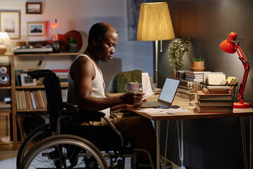Mature african american man in wheelchair sitting at desk in his bedroom and looking through bills