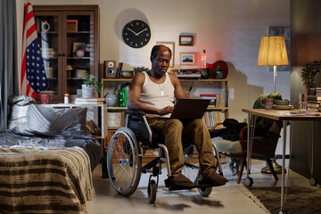 Black man with physical disability sitting in his bedroom and browsing laptop working remotely