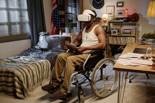 Black man in wheelchair wearing virtual reality headset and holding digital tablet sitting at home