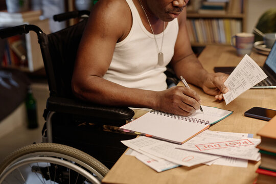 Unrecognizable black man in wheelchair writing information from piece of paper into his notebook