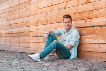 Handsome fashionable guy with a hairstyle in stylish summer casual clothes with a shirt, jeans and sneakers sits near a wooden wall on the street