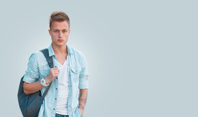 Stylish handsome young man with hairstyle in a fashion denim shirt with a backpack on a blue...