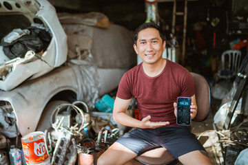 Handsome young mechanic shows a cell phone screen sitting on a chair in an old garage