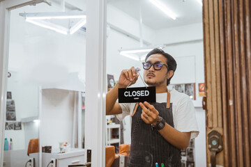 handsome asian barber wearing apron holding closed sign at barbershop