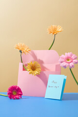Fresh gerbera flowers are decorated around a pink envelope, next to a blue card with the words HAPPY WOMEN'S DAY. Women's day theme with copy space for ads.