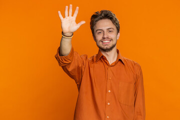 Young Caucasian man smiling friendly at camera, waving hands gesturing hello greeting or goodbye welcoming with invitation hospitable expression. Handsome guy isolated on orange studio background - obrazy, fototapety, plakaty