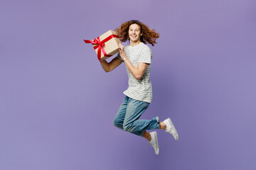 Full body young man wears grey striped t-shirt casual clothes hold in hand present box with gift ribbon bow jump high look aside isolated on plain pastel purple background studio. Lifestyle concept. - obrazy, fototapety, plakaty