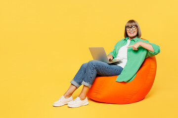 Fototapeta na wymiar Full body fun elderly IT woman 50s years old wear green shirt glasses casual clothes sit in bag chair hold use work point on laptop pc computer isolated on plain yellow background. Lifestyle concept.