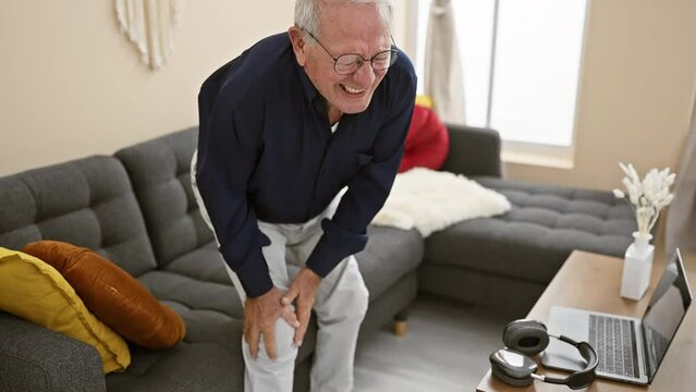 Unhappy senior man suffering knee pain, sitting alone on sofa indoor at home, sporting grey hair, glasses and worry expression amid living room interior