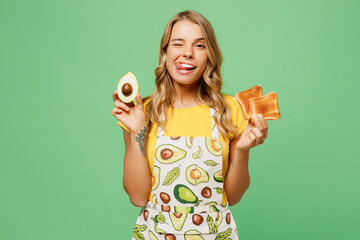 Young fun housewife housekeeper chef cook baker woman wear apron yellow t-shirt hold in hand avocado, bread toast wink blink eye isolated on plain pastel green background studio. Cooking food concept.