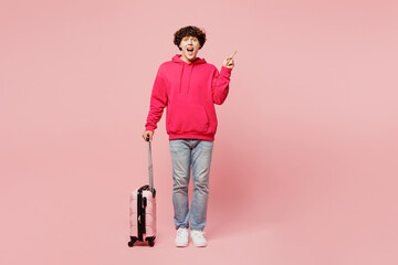 Traveler man wear hoody casual clothes hold suitcase bag point finger aside isolated on plain pink...