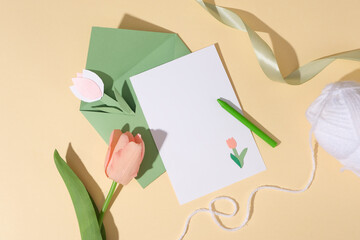 Flat lay of a white paper and green envelope decorated with a crayon, ribbon and a white skein. A tulip flower and paper tulip flowers featured. Copy space on the card for content about Women’s Day