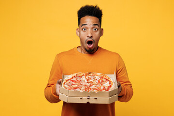 Young surprised man wear orange sweatshirt casual clothes holding Italian pizza in cardboard flatbox isolated on plain yellow background. Proper nutrition healthy fast food unhealthy choice concept.