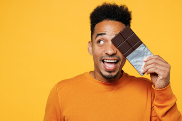 Young fun man wear orange sweatshirt casual clothes hold eat bar of chocolate look aside on area...