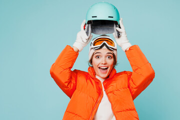 Young surprised shocked skier woman wear warm padded windbreaker jacket hat ski goggles mask hold put on helmet travel rest spend weekend winter season in mountains isolated on plain blue background.
