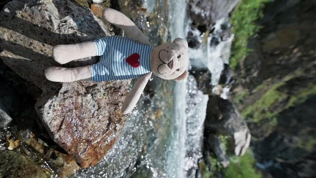 toy plush teddy bear sits on a rock by a mountain river on a camping trip in summer. Vertical slow motion