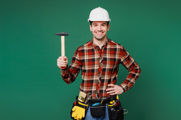 Young fun employee handyman man wear red shirt hardhat hat work hold hammer stand akimbo isolated...