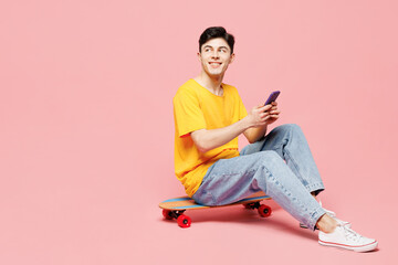 Full body young fun man he wears yellow t-shirt casual clothes sit on skateboard hold in hand use mobile cell phone isolated on plain pastel light pink background studio portrait. Lifestyle concept.