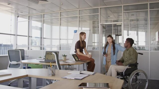 Wide copy space shot of young African American man in wheelchair and his two Caucasian female colleagues having conversation in glass walled office during coffee break