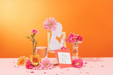 Gerberas, carnations and glassware are displayed around a women's day card on an orange background with a pink surface. Copy space with front view.
