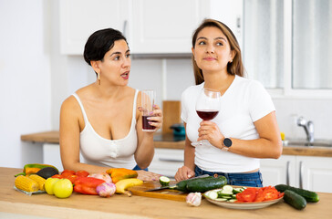 Two cheerful young Hispanic female roommates enjoying weekend together, drinking wine and preparing vegetable dish for dinner at home kitchen