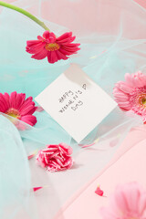 A white card with the words Happy Women's Day is placed next to bright fresh flowers. A pastel blue mesh fabric decorated on a pink background.