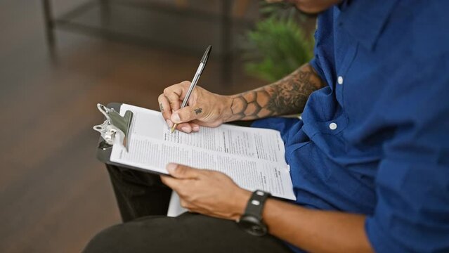 Concentrated hispanic man with tattooed hands sitting in chair, seriously writing on clipboard in roomy business waiting room, diligently performing his indoor work