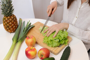 Unrecognizable unknown woman sitting at kitchen table with fruit and vegetables cutting green lettuce leaves for healthy salad
