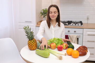 Healthy lifestyle. Raw vegan. Domestic kitchen. Nutritious dinner. Caucasian brown haired woman sitting at kitchen table with fruit and vegetables cooking organic meals at home