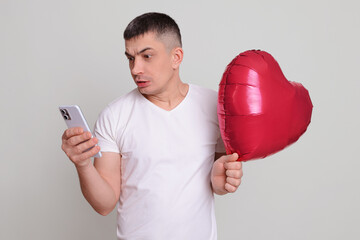 Confused shocked Caucasian brunette man holding red helium balloon in heart shape isolated over...