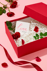 Close-up of the inside of a gift box with red roses, rose leaves and a greeting card. Silk ribbons and fresh roses on a pastel pink background.