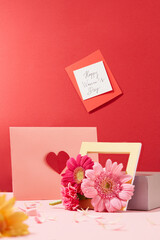 A note with the words happy women's day stuck on a red background. A wooden picture frame, a greeting card and fresh flowers are displayed with a women's day theme.