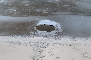 Hole with water in ice on the surface of a frozen river near the shore on a winter day